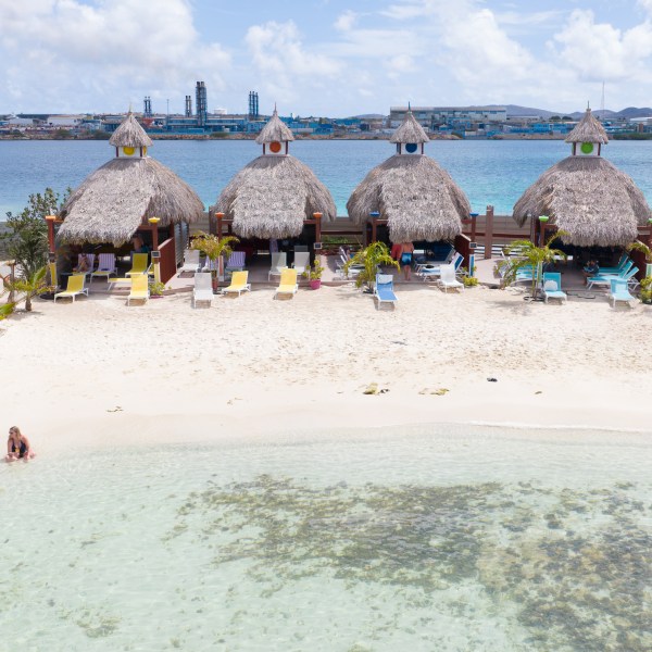Beach with straw huts, lounge chairs, two people by water, industrial buildings in background.