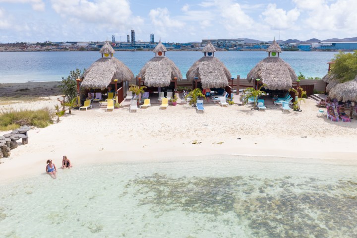 Beach with straw huts, lounge chairs, two people by water, industrial buildings in background.
