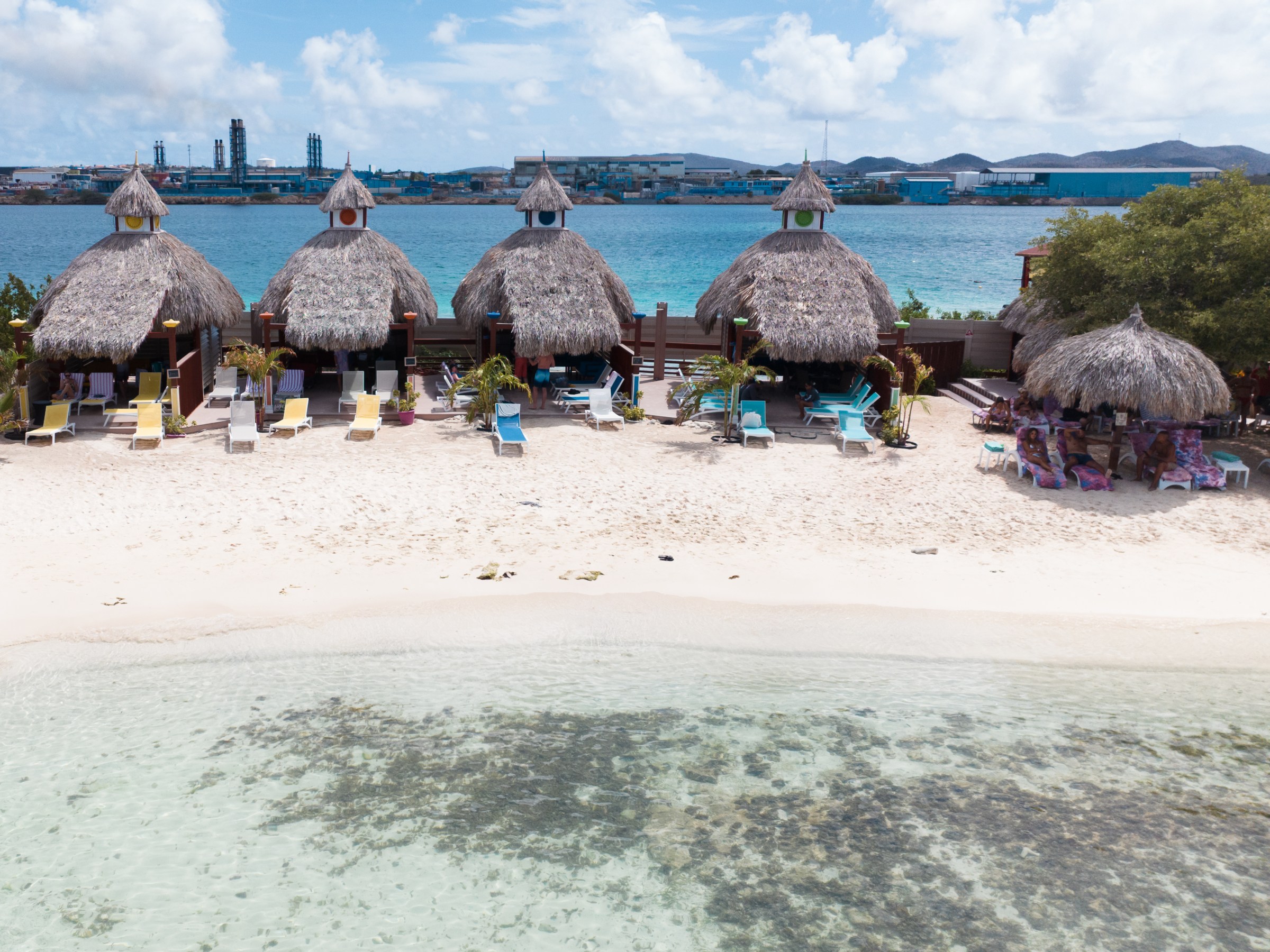 Rows of thatched huts on a sandy beach with chairs, facing a blue ocean.
