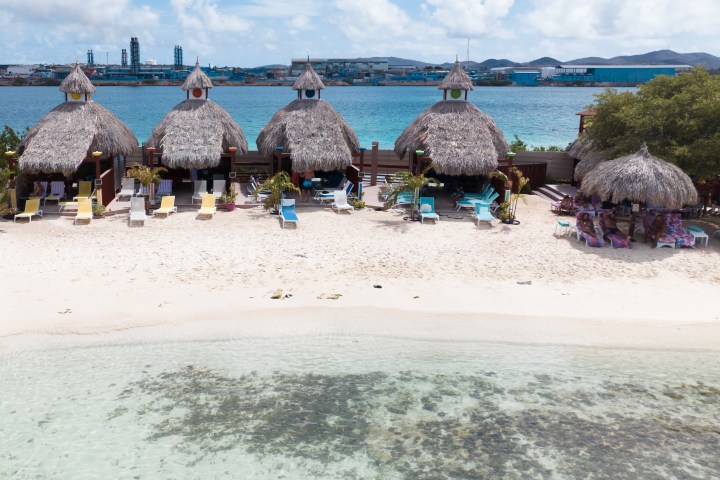 Rows of thatched huts on a sandy beach with chairs, facing a blue ocean.