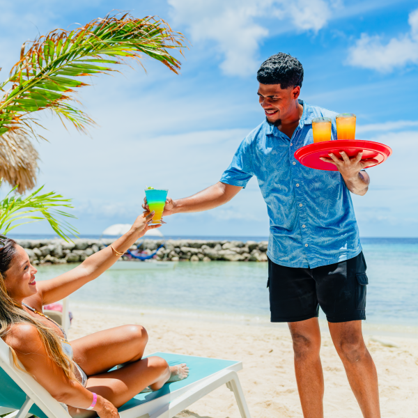 Woman on beach chair receiving drink from server with tray, ocean in background.