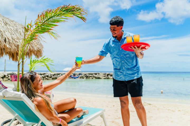 Woman on beach chair receiving drink from server with tray, ocean in background.