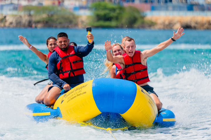 Four people in red life jackets riding a yellow inflatable banana boat on the ocean.