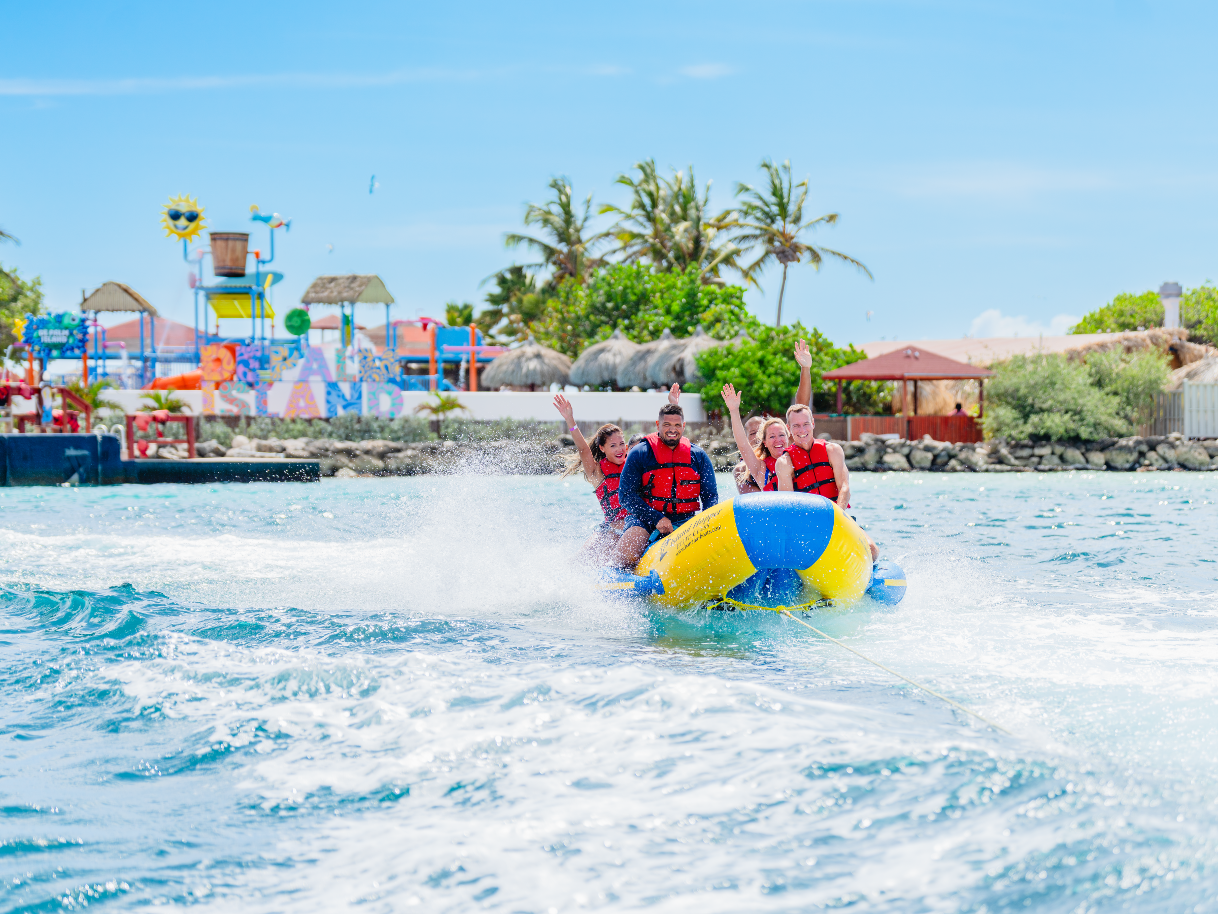 Group wearing life jackets on an inflatable banana boat in the ocean, amusement park in the background.