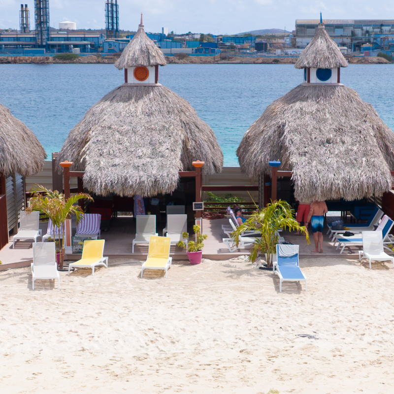 Four thatched-roof huts with colorful chairs on a sandy beach near water.