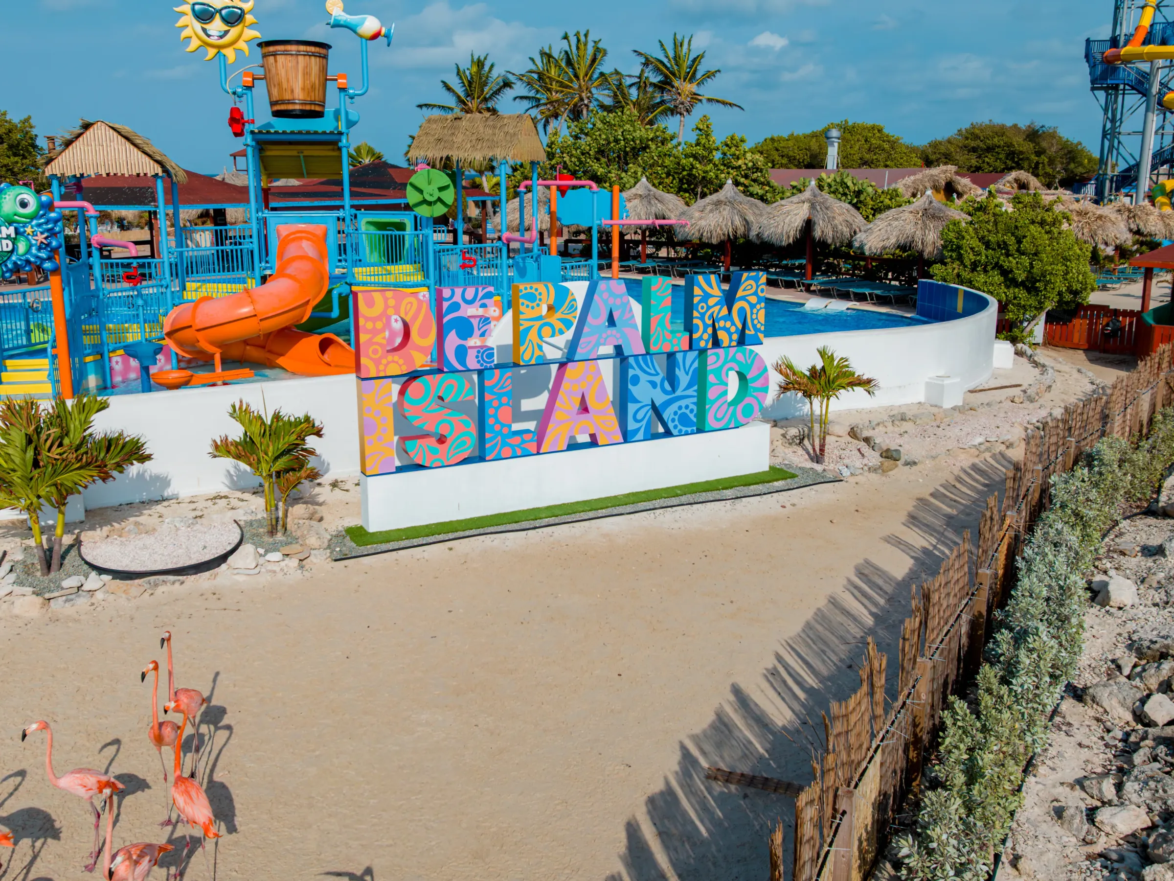 Colorful water park with a palm island sign and flamingos on a sandy path.