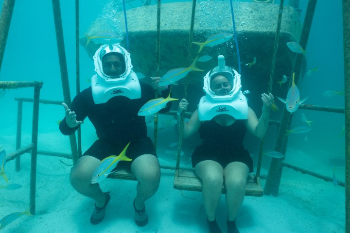 Two people underwater sitting on swings, wearing helmets, with fish swimming around them.