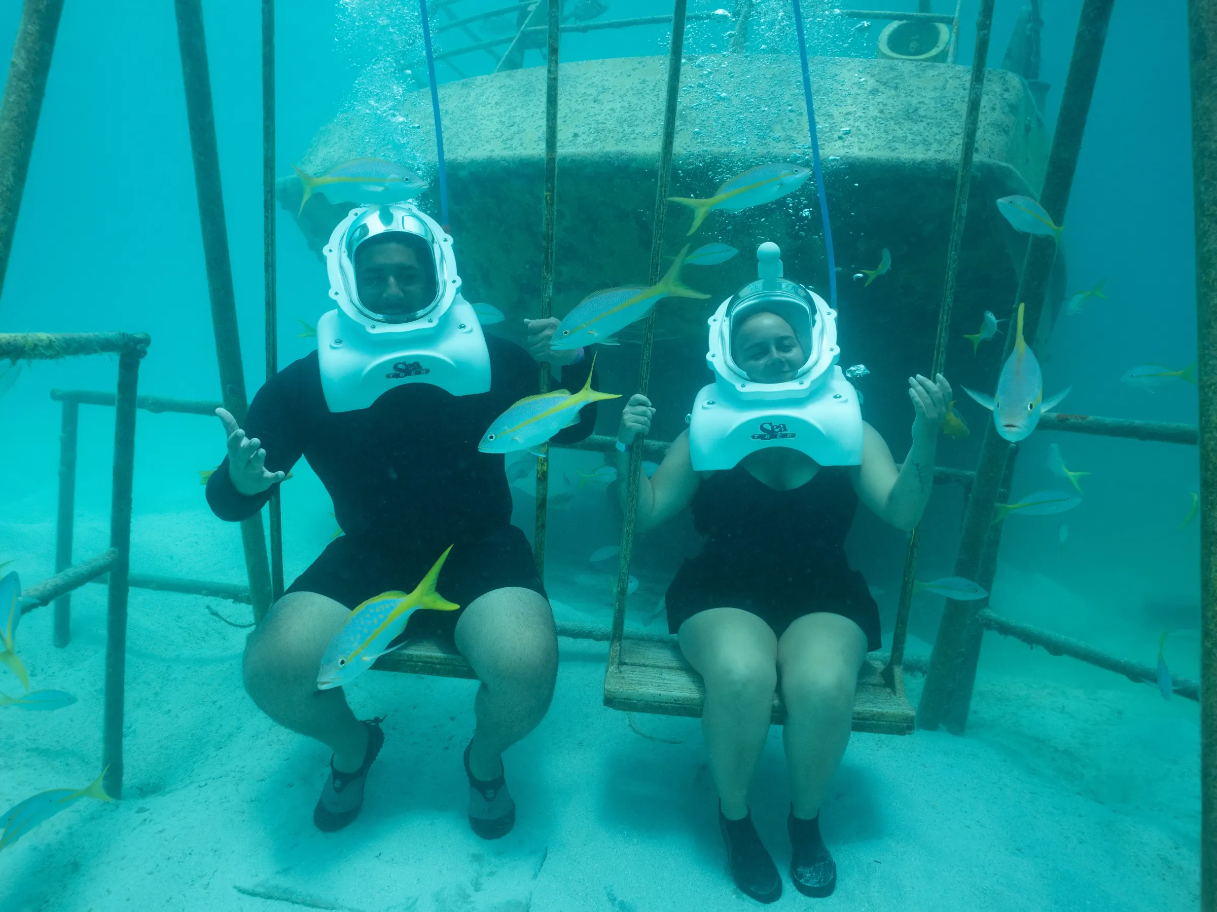 Two people underwater wearing helmets, sitting on swings, surrounded by fish.