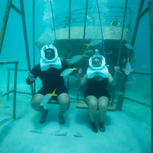 Two people underwater wearing helmets, sitting on swings, surrounded by fish.