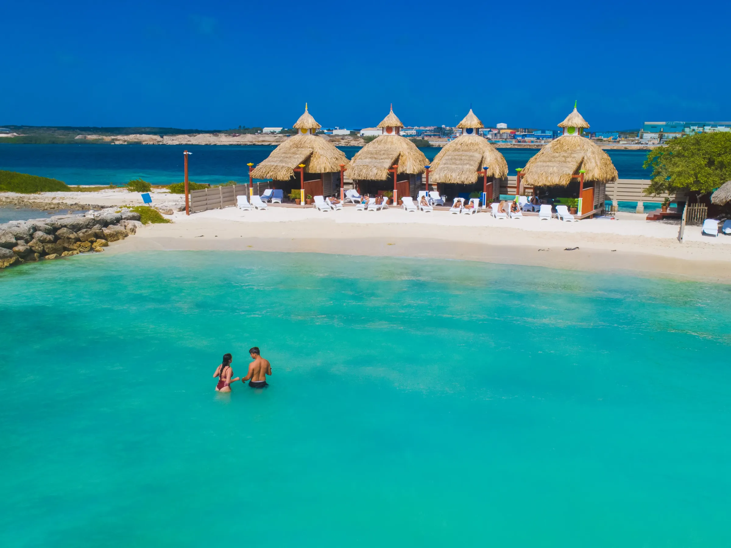 Couple wading in turquoise water near beach huts on a sunny day.