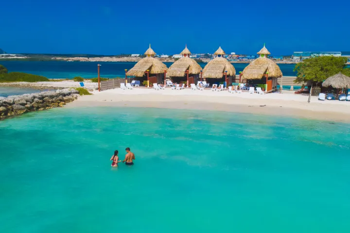 Couple wading in turquoise water near beach huts on a sunny day.