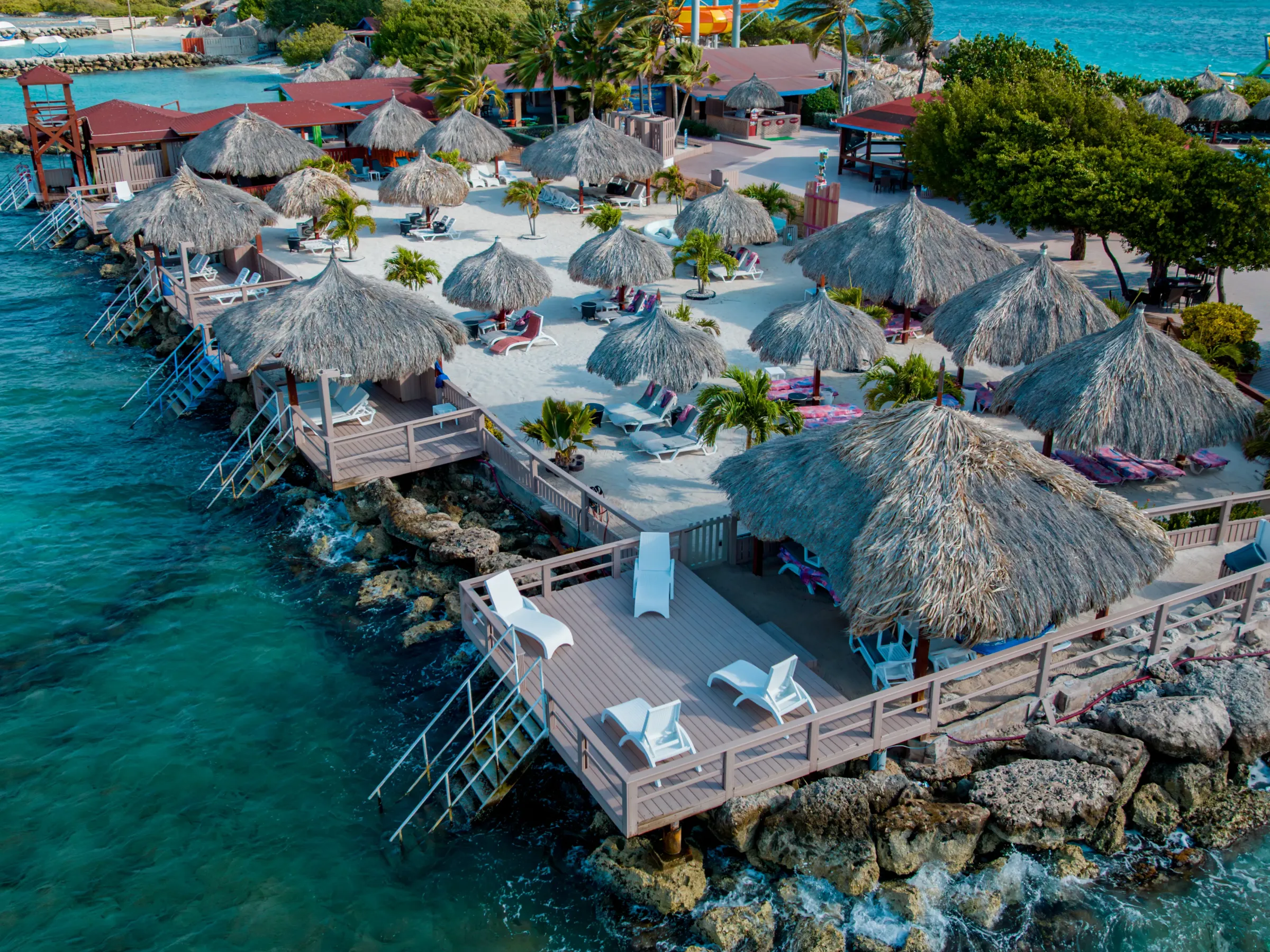 Island resort with straw huts by turquoise water and rocky shore.